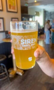Craft beer glass featuring Siren Craft Brew logo, in a lively brewery setting.