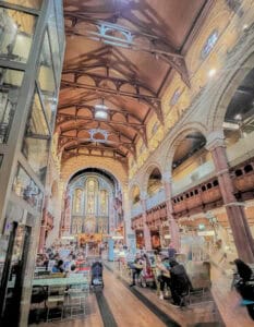 Library interior with high vaulted ceiling and colorful stained glass windows, lively cafe atmosphere, and people enjoying drinks and conversations.
