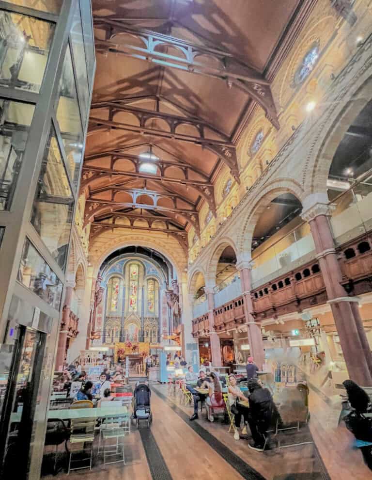 Library interior with high vaulted ceiling and colorful stained glass windows, lively cafe atmosphere, and people enjoying drinks and conversations.